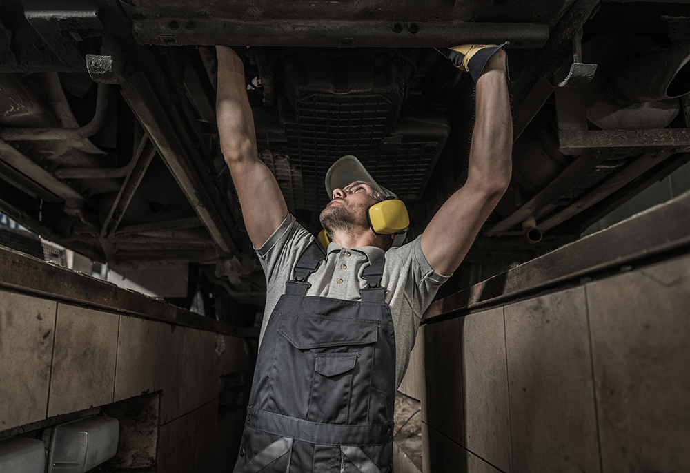 Caucasian Automotive Mechanic Under Coach Bus Looking For Potential Issue with the Vehicle Diesel Engine. Transportation Industry Theme.